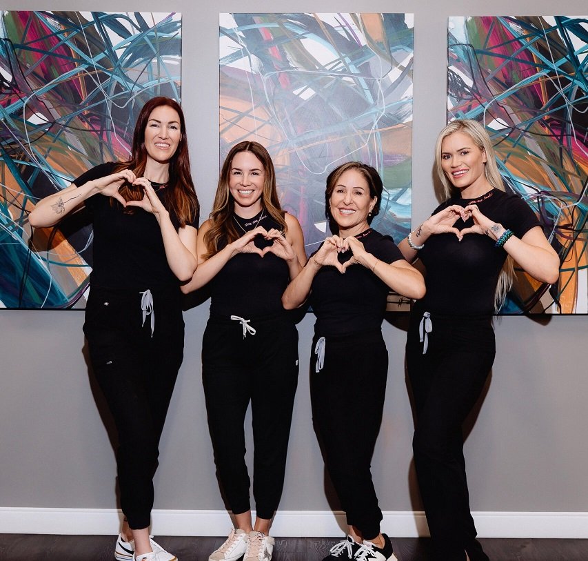 Four women in black outfits making heart shapes with their hands in front of abstract art.