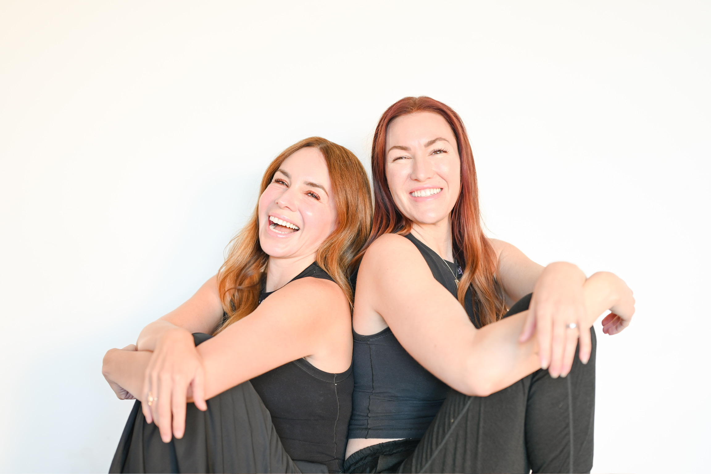 Two smiling women sitting back-to-back in black tops against a white background