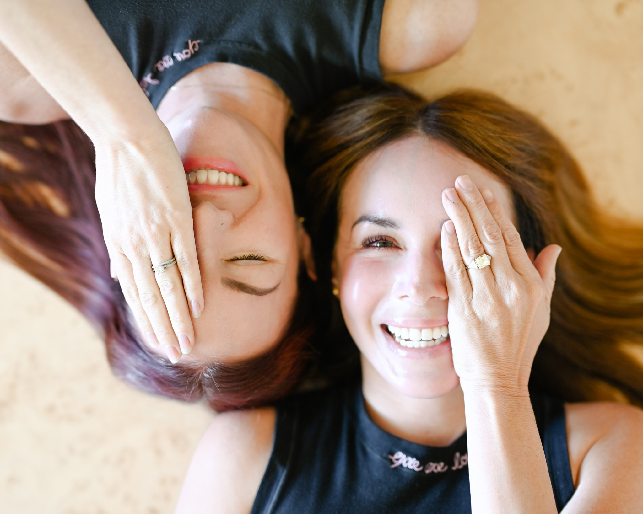 Two smiling women lying on the floor head-to-head, each covering one eye with a hand