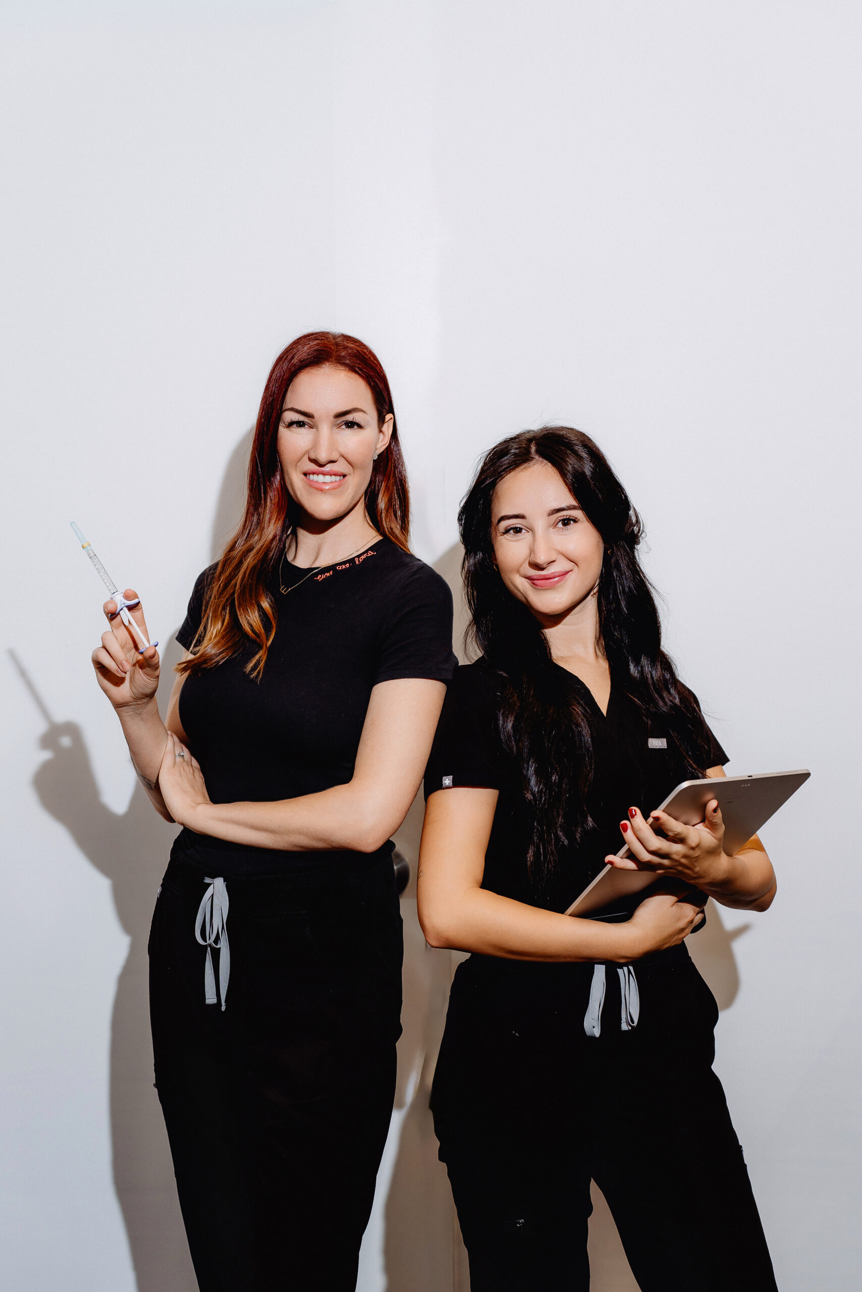 Two Be You Medical nurses in black uniforms, one with a Restylane filler syringe and one with a tablet